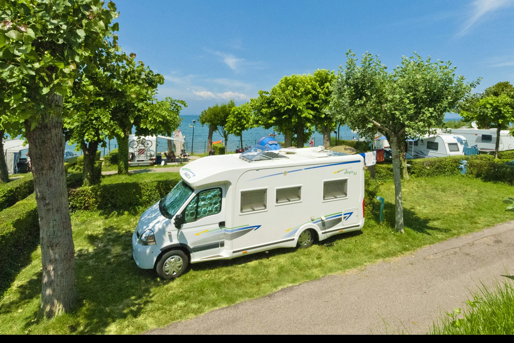 Wohnmobil auf einem Campingplatz-Stellplatz, neben Bäumen und Hecken, mit Rasenfläche und Kiesweg im Hintergrund.