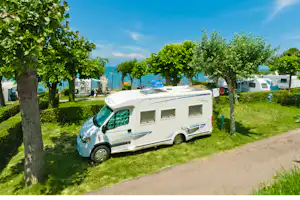 Wohnmobil auf einem Campingplatz-Stellplatz, neben Bäumen und Hecken, mit Rasenfläche und Kiesweg im Hintergrund.