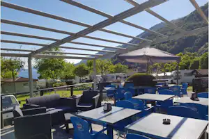 Überdachte Camping-Terrasse mit blauen Stühlen und Tischen, Sonnenschirmen, Hecken und Blick auf grüne Hügel.