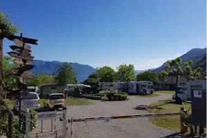 Einfahrt eines Campingplatzes mit Wohnmobilen und Autos, Kiesweg, Bäumen und Bergen im Hintergrund.