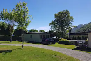 Campingplatz mit Wohnwagen-Stellplatz, Kiesweg und Rasenfläche vor einem modernen Gebäude, daneben ein Auto und Bäume.