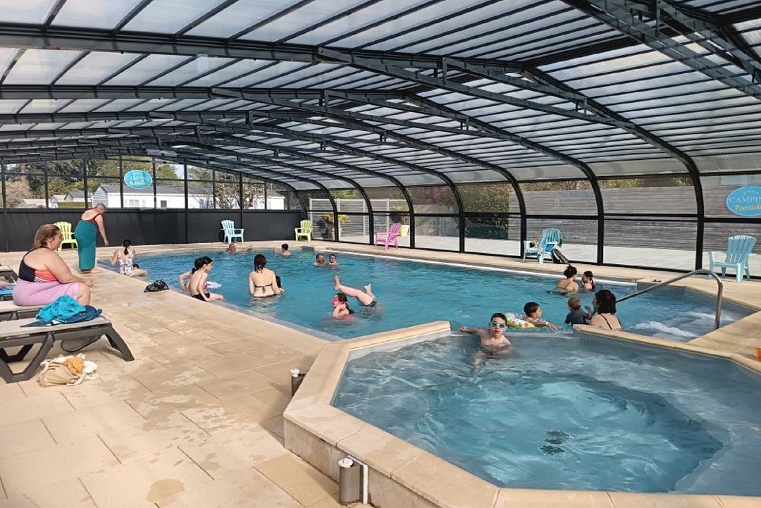 Überdachtes Schwimmbecken auf dem Campingplatz, Menschen im Wasser, Liegen und Stühle am Beckenrand.