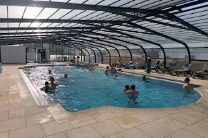 Innenpool mit blauem Wasser, mehreren Liegen und Sonnenschirmen unter einem Glasdach, im Hintergrund ein Zaun und Gebäude.