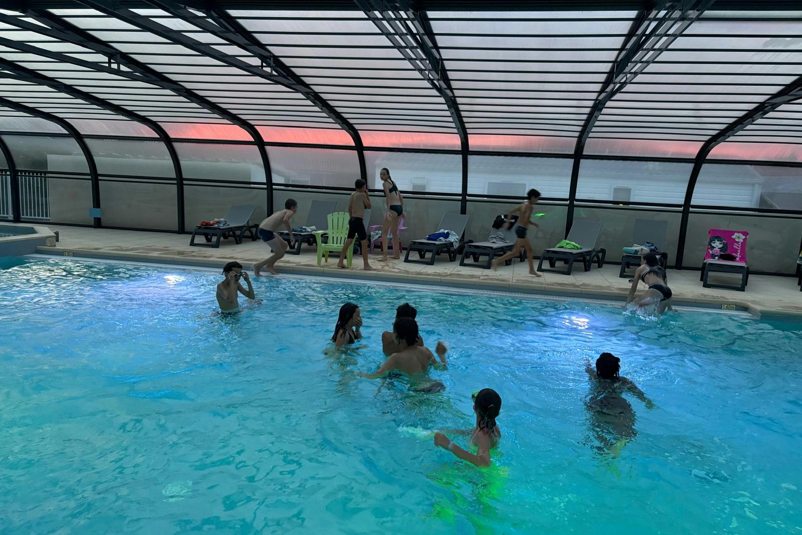 Hallenbad mit blauem Schwimmbecken, mehreren Liegen und Sonnenschirmen unter einem Glasdach.