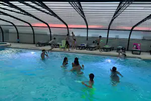 Hallenbad mit blauem Schwimmbecken, mehreren Liegen und Sonnenschirmen unter einem Glasdach.