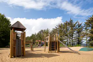 Holz-Spielturm mit Dach, Rutsche und Seilnetz auf dem Sandspielplatz, umgeben von Bäumen und Blick auf Wasser.
