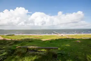 Blick auf den Strand mit Steg, Sandweg und grüner Wiese im Vordergrund, dahinter das Meer und große Wolken.