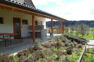 Ferienhaus mit überdachter Terrasse, Grill und Gartenbeet mit Sträuchern vor dem Gebäude.