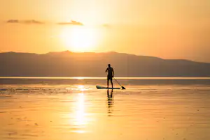 Paddler auf einem Stand-Up-Paddleboard im See, im Hintergrund Berge und die spiegelnde Abendsonne.