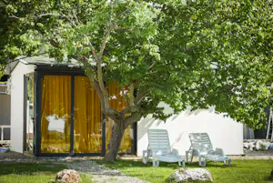 Stellplatz mit Wohnwagen und Terrasse, weiße Fassade, gelbe Vorhänge, zwei Kunststoffliegen unter einem großen Baum.