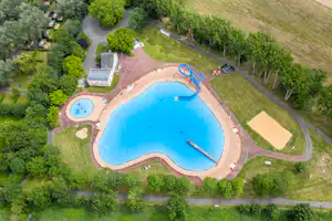 Luftaufnahme eines Campingplatzes mit großem blauem Schwimmbecken, zwei kleineren Becken, Liegeflächen und Wegen im Grünen.