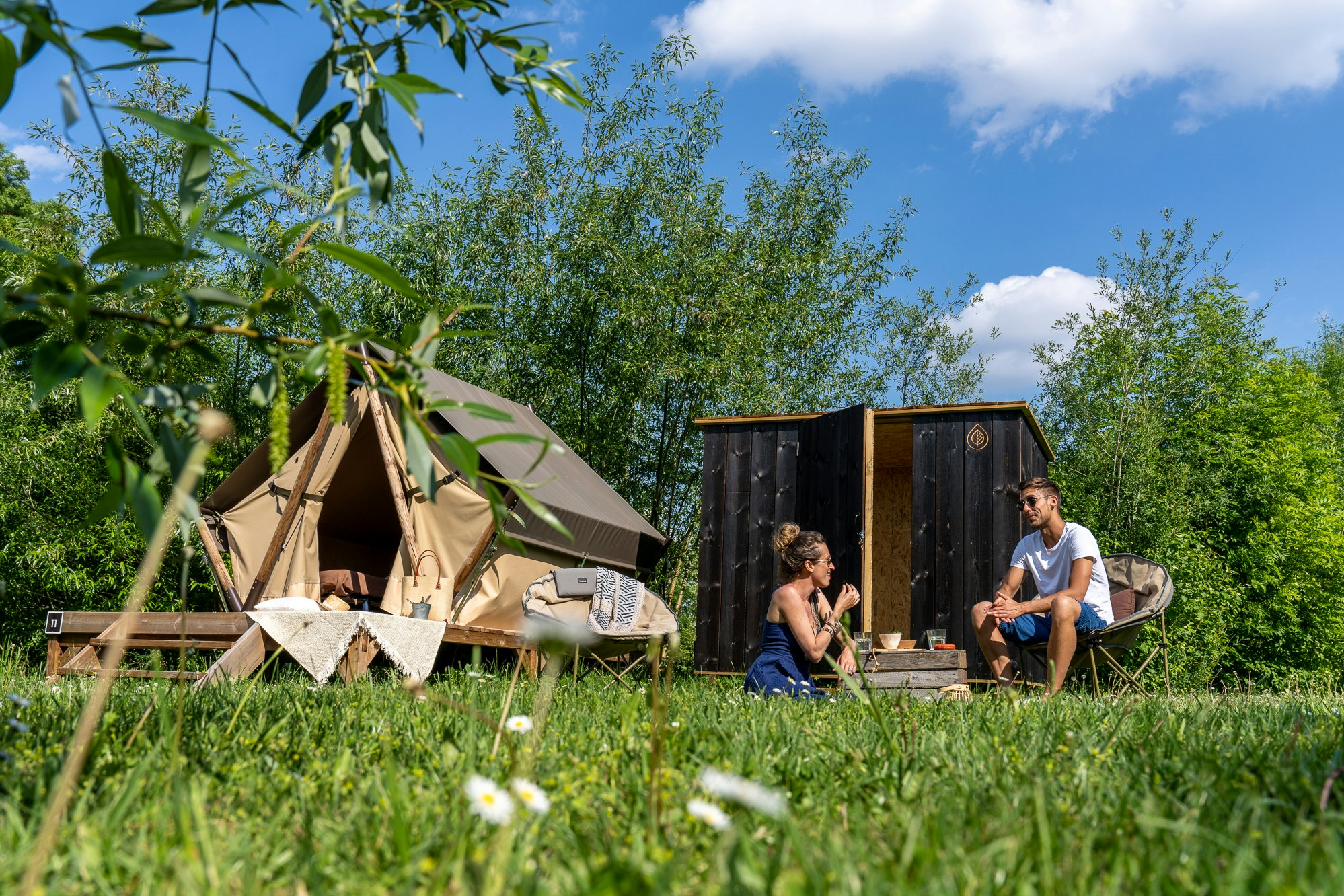 Zelt und Wohnwagen auf einem Campingplatz im Grünen, im Vordergrund Gras und Blumen, daneben zwei Personen am Tisch.