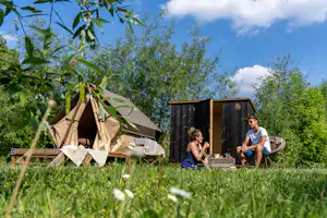 Zelt und Wohnwagen auf einem Campingplatz im Grünen, im Vordergrund Gras und Blumen, daneben zwei Personen am Tisch.