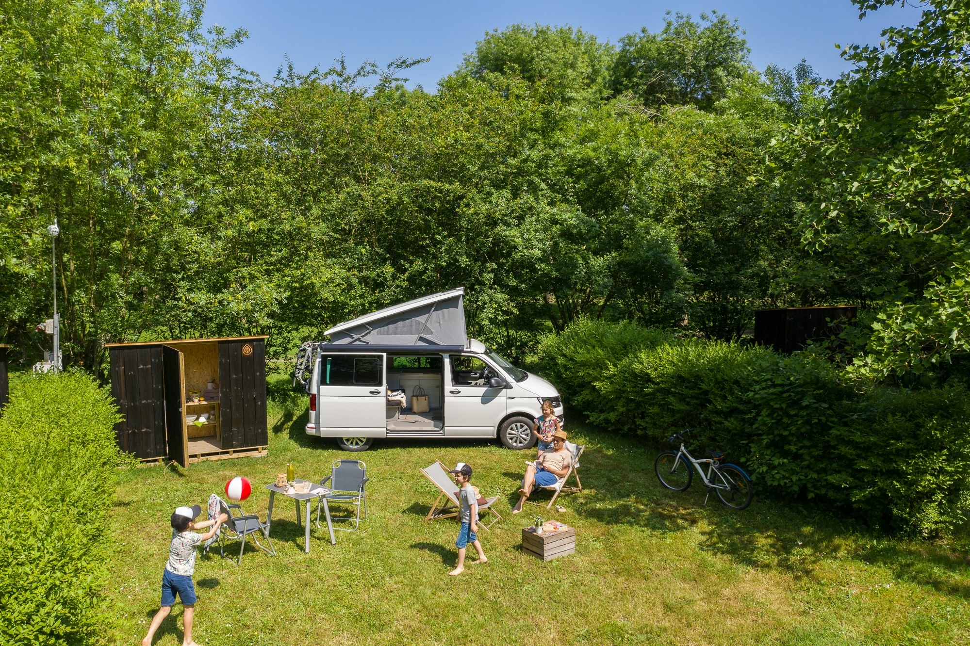 Wohnmobil auf einem Campingplatz im Grünen, daneben Spielzeug und Kinder auf dem Rasen, Hecke und Bäume im Hintergrund.