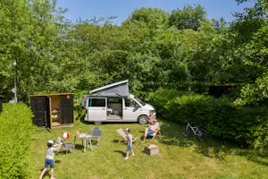 Wohnmobil auf einem Campingplatz im Grünen, daneben Spielzeug und Kinder auf dem Rasen, Hecke und Bäume im Hintergrund.