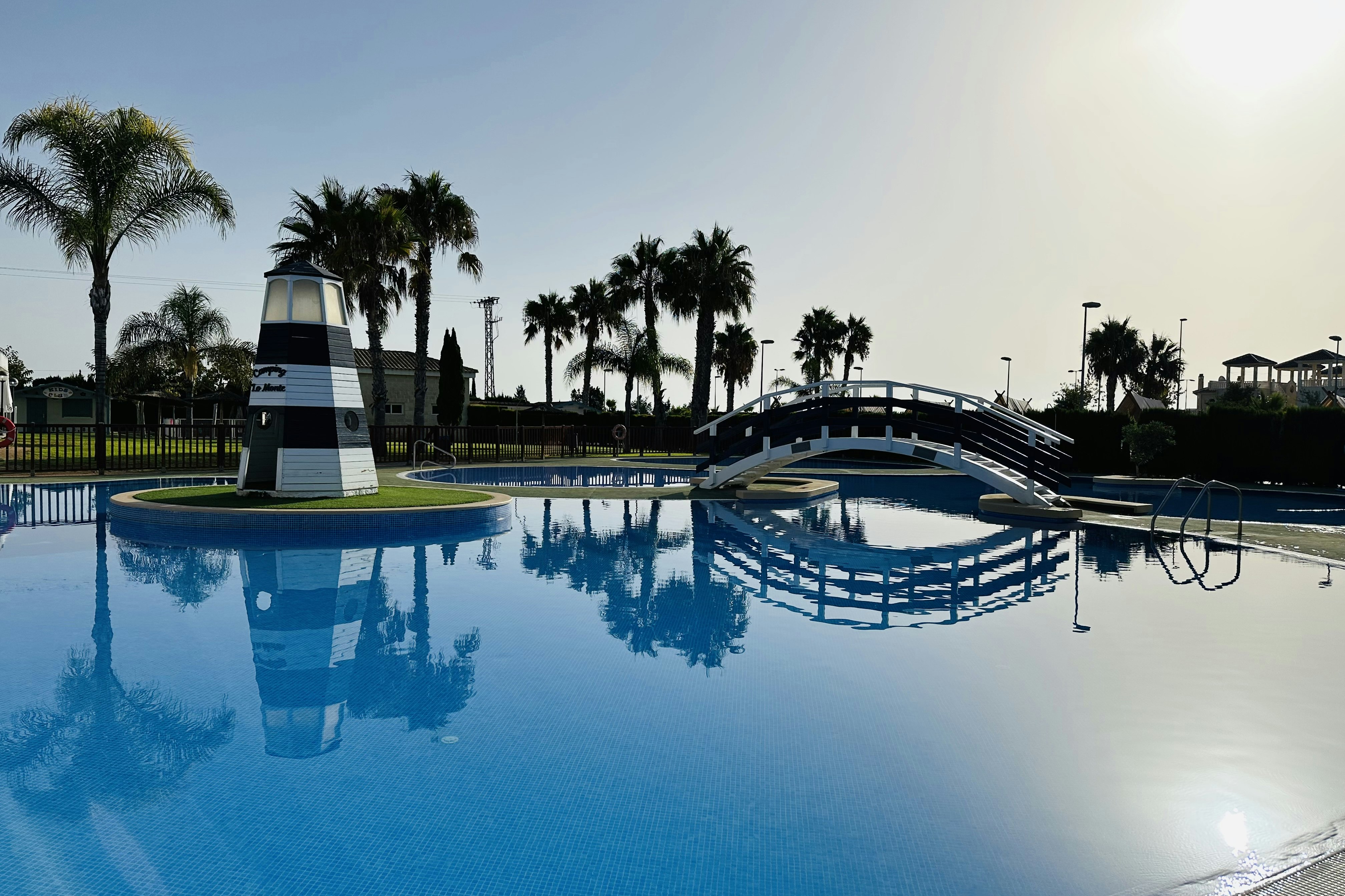 Großer Camping-Pool mit Liegen und Sonnenschirmen, Palmen im Hintergrund und Blick auf das Poolgeländer.