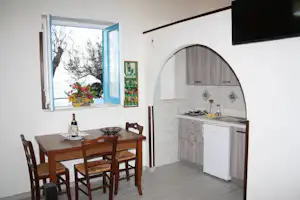 Kleine Küchenzeile mit Gasherd und Spüle, daneben Esstisch mit Stühlen und Fensterblick ins Freie.