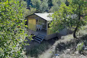 Holzhütte mit überdachter Terrasse und zwei Stühlen, umgeben von Bäumen und Sträuchern im grünen Hanggelände.