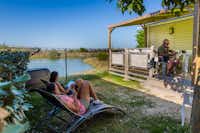 Camping L'Océan - Gäste an den Liegestühlen und auf der Terrasse vor dem Mobilheim mit dem Blick auf Wasser