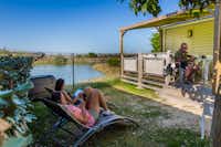 Camping L'Océan - Gäste an den Liegestühlen und auf der Terrasse vor dem Mobilheim mit dem Blick auf Wasser