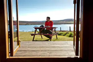 Mann am Tisch auf einer Holzterrasse mit Blick auf einen See, eingerahmt durch offene Tür und Fenster.
