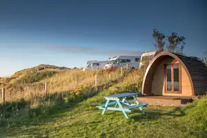 Holzkuppeltent auf einer Wiese mit zwei Liegestühlen, im Hintergrund Wohnwagen und Wohnmobil vor Hügeln.
