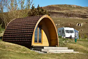 Holzhütte mit Fenster und Satteldach auf einem Campingplatz, im Hintergrund Wohnmobil und Hügel mit Bäumen.