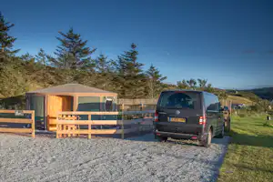Wohnwagen und Transporter auf einem Campingplatz mit Holzgeländer, Kiesfläche und Wiese, im Hintergrund Bäume und Hügel.
