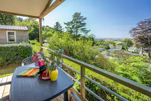 Blick von der Terrasse auf einen Bungalow mit Garten, Blumen auf dem Tisch und Häusern im Tal, umgeben von Bäumen.