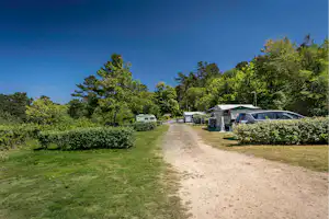 Campingplatzweg zwischen Stellplätzen mit Wohnwagen und Auto, umgeben von Hecken und Rasen, unter blauem Himmel.