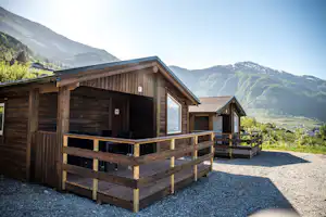 Holzhütte mit Terrasse, Sitzgelegenheiten und großen Fenstern, im Hintergrund Berge und ein Tal mit Häusern.