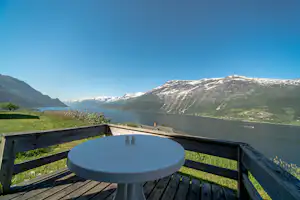 Blick von der Terrasse auf einen See und schneebedeckte Berge, mit Holzzaun und rundem Tisch im Vordergrund.
