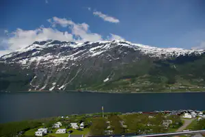 Campingplatz am See mit Wohnmobilen und Zelten, Blick auf schneebedeckte Alpen und bewaldete Hänge unter blauem Himmel.