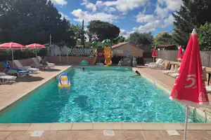 Schwimmbecken auf dem Campingplatz mit Liegen, roten Sonnenschirmen, Spielzeugboot im Wasser und Bäumen im Hintergrund.