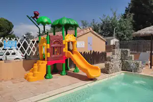 Bunter Spielplatz mit Rutsche und Kletterelementen neben dem Campingpool, umgeben von Steinboden und Hecken.