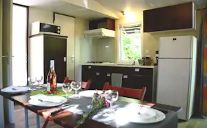 Wohnküche mit Esstisch, Küchenzeile mit Kühlschrank und Herd, Fenster mit Rollladen und Sitzbank im Hintergrund.
