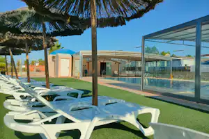 Weiße Liegen unter einem Sonnenschirm am Campingplatz, daneben ein umzäunter Pool mit Glasüberdachung und Rasenfläche.