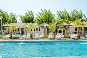 Blauer Pool mit Liegestühlen am Rand, Palmen und Sonnenschirme vor weißen Gebäuden mit grünen Fensterläden.