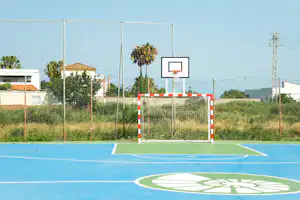 Handballtor auf einem Sportplatz mit blauem Boden, Netz und Umzäunung, im Hintergrund Häuser und Bäume.