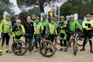 Radteam in grünen Trikots mit Helmen und Fahrrädern auf einem Campingplatzweg, im Hintergrund Bäume und Flaggen.
