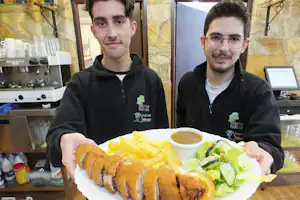 Zwei Personen servieren ein Teller mit Pommes, Salat und Bratwurst in einer Camping-Kantine mit Kaffeemaschine im Hintergrund.