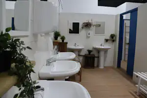Camping-Badezimmer mit mehreren Waschbecken, Holzablagen und Holzboden, weiße Fliesenwand und blaue Tür im Hintergrund.