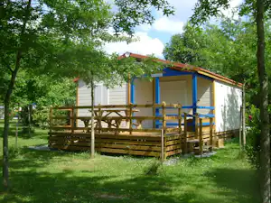 Mobilheim auf Campingplatz mit Holzterrasse, weißen Wänden und blauem Rahmen, umgeben von Bäumen und Rasen.