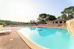 Schwimmbad mit blauem Wasser, Randsteinen und mehreren Liegen am Campingplatz, im Hintergrund Gebäude und Bäume.