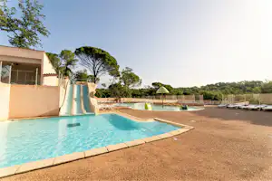 Schwimmbecken mit Poolrand und Liegen am Campingplatz, daneben ein Gebäude und Bäume im Hintergrund.