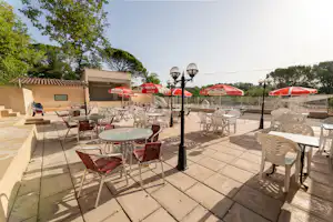Terrasse mit Tischen und Stühlen, roten Sonnenschirmen und Laternen vor einem Gebäude, umgeben von Bäumen.