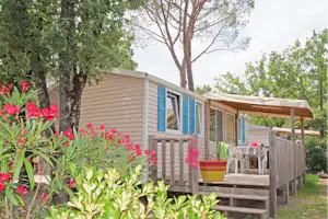 Bungalow mit Veranda und Holzgeländer, davor Blumenbeete und Rasen, im Hintergrund Bäume und ein weiterer Wohnwagen.