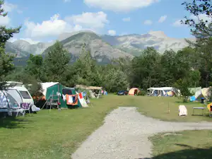 Campingwiese mit mehreren Zelten, einem grünen Zelt und Wohnwagen, im Hintergrund Berge und Bäume.