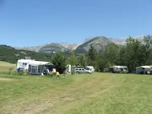 Campingplatz auf grünem Rasen mit Wohnwagen, Zelt und Picknicktisch, im Hintergrund Berge und Bäume.