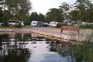 Teich mit Ufersteinen und Schilf, dahinter Stellplätze mit Wohnwagen und Wohnmobilen zwischen Bäumen.
